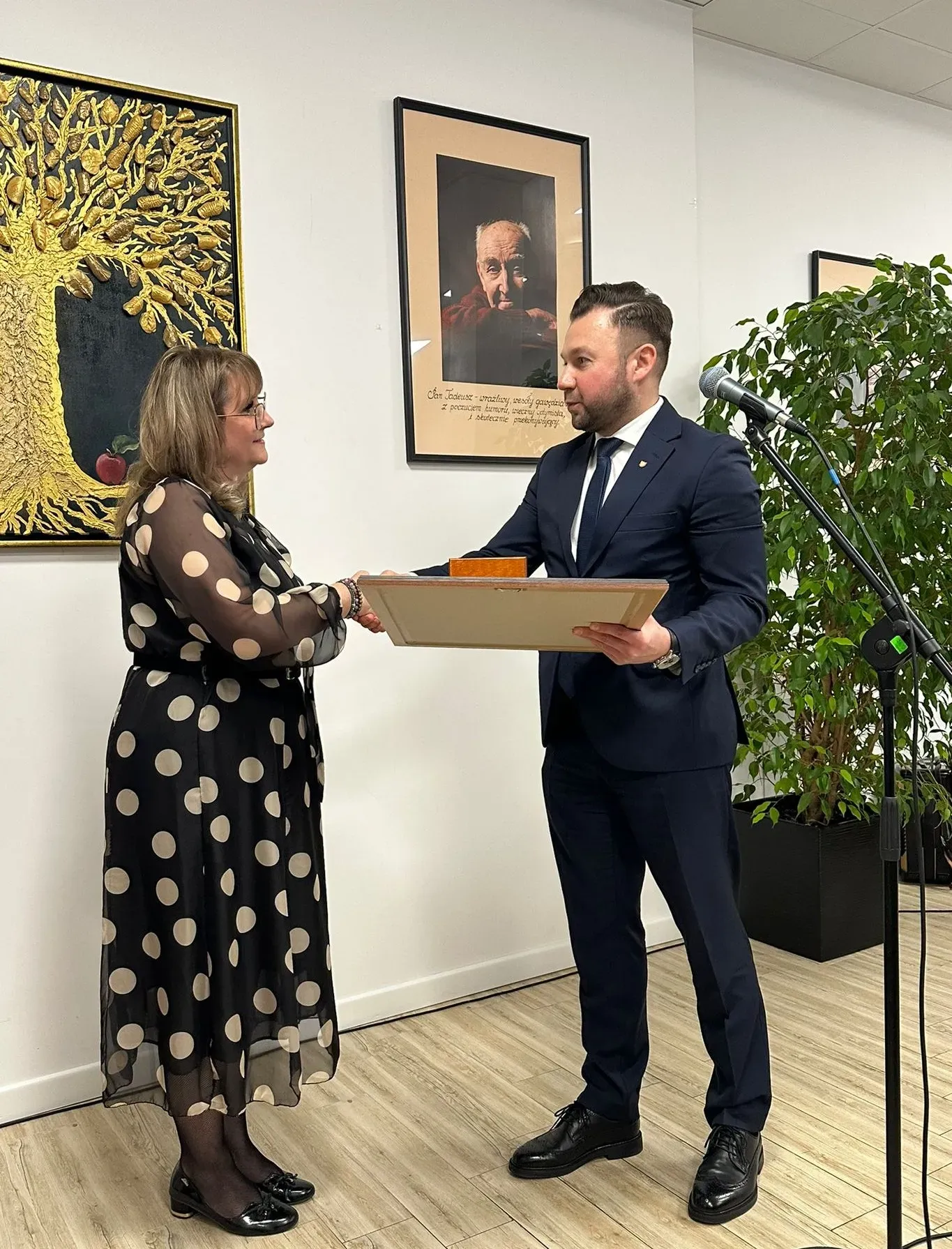 Jasne pomieszczenie. Mężczyzna w granatowym garniturze przekazuje kobiecie w czarnej sukience w beżowe grochy ramkę z wyróżnieniem, stojąc na jasnej podłodze. W tle na ścianie wiszą dwa obrazy, po prawej stronie stoi mikrofon na statywie i wysoka zielona roślina w doniczce.